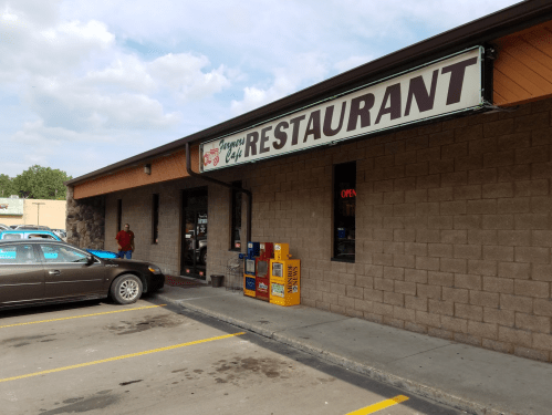 Exterior of a restaurant with a sign reading "Restaurant," featuring a parked car and a newspaper stand outside.