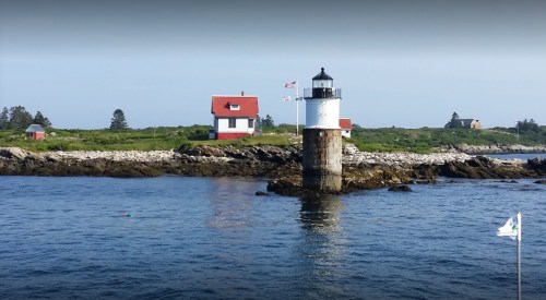 A lighthouse on a rocky shore with a red-roofed house nearby, surrounded by calm blue water and greenery.