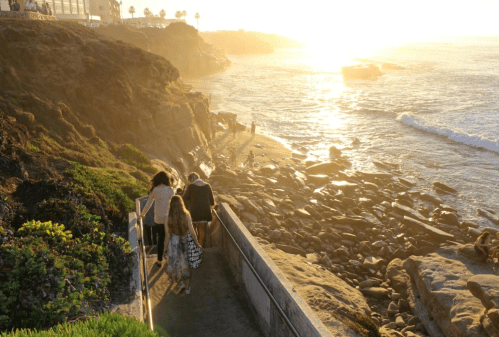 A family walks down a path towards a rocky beach at sunset, with golden light reflecting on the water.