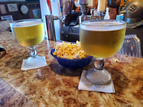 Two large glasses of beer sit on a bar counter next to a blue bowl of popcorn.