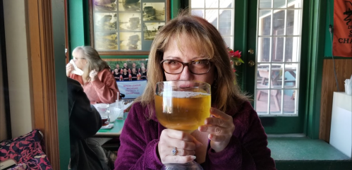 A woman with glasses holds a large glass of beer, smiling at the camera in a cozy setting.