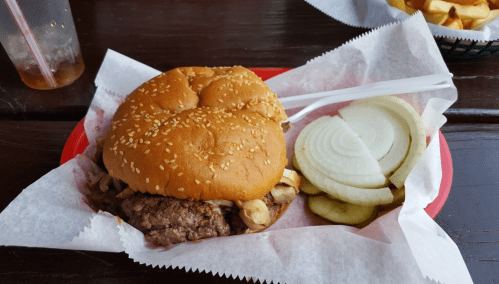 A sesame seed bun burger with mushrooms, served with sliced onions and pickles on a red plate.