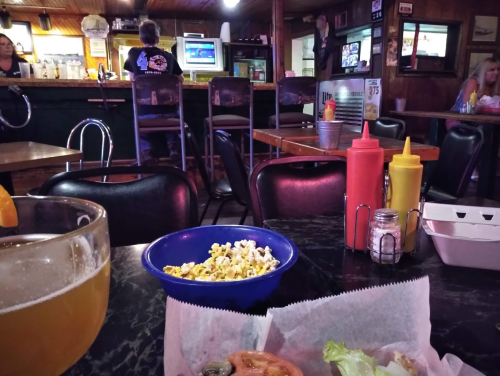 A cozy bar scene with a bowl of popcorn, a drink, and condiments on a table, with patrons in the background.