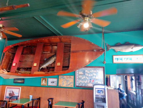 Interior of a restaurant featuring a wooden boat on the wall, fish decor, and a glimpse of a dining area.