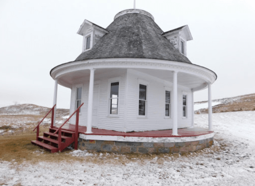 A round, white building with a gray shingled roof and red steps, set in a snowy landscape.