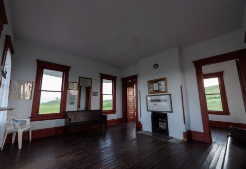 A spacious room with wooden floors, red trim, mirrors, and a fireplace, featuring large windows and a simple chair.
