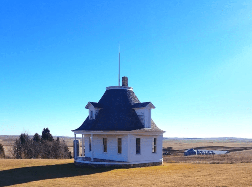 A white, octagonal house with a dark shingle roof stands on a grassy hill under a clear blue sky.