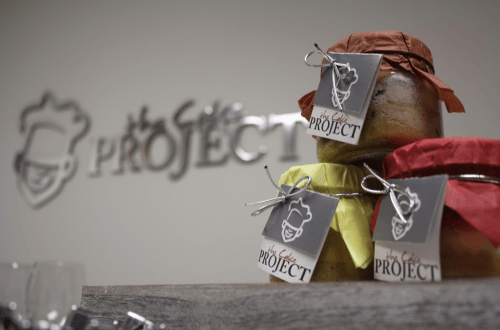 Three jars with colorful lids and tags sit in front of a sign that reads "The Cake Project."