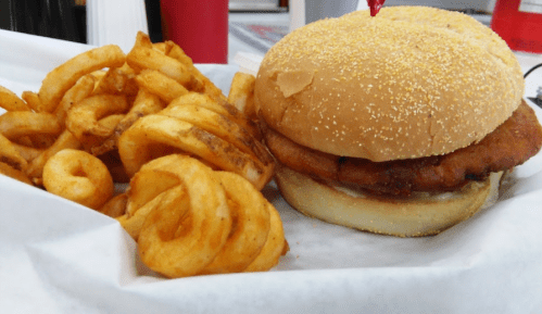 A crispy chicken sandwich on a bun with a cherry on top, served with curly fries on the side.