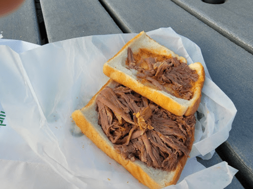 A sandwich with shredded beef on two slices of white bread, resting on a paper wrapper on a wooden table.