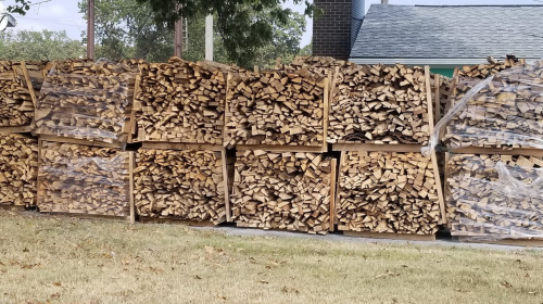Stacked firewood neatly arranged in wooden crates, with some wrapped in plastic, set against a grassy area.