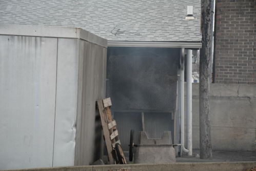 Smoke billows from a narrow alley between a shed and a building, with a wooden pallet and a cart visible.