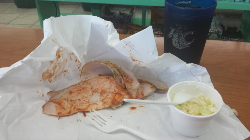 A partially eaten sandwich with turkey and sauce on a paper wrapper, alongside a cup of coleslaw and a drink.