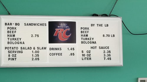 Menu board displaying prices for BBQ sandwiches, drinks, and sides, including pork, beef, turkey, and bologna options.