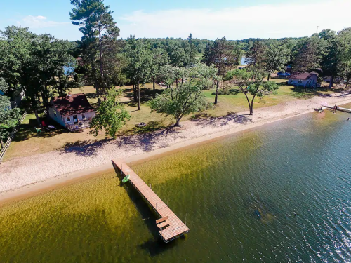 Aerial view of a sandy beach with a wooden dock, surrounded by trees and a calm lake on a sunny day.