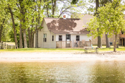 A cozy lakeside cottage surrounded by trees, with a sandy beach and a small deck overlooking the water.