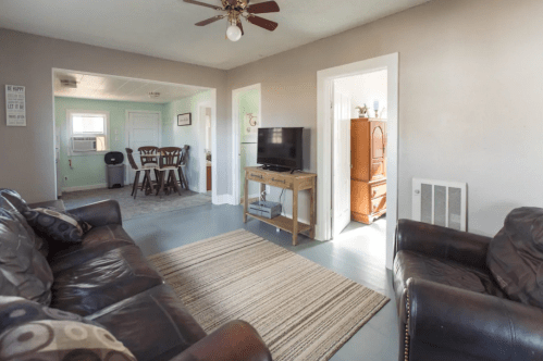 Cozy living room with a sofa, armchair, TV, and a rug, leading to a dining area and a bright room beyond.