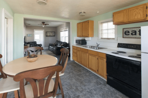 A cozy kitchen with wooden cabinets, a round table, and a view into a living room with black sofas.