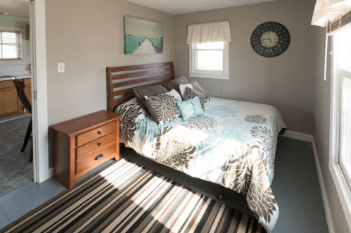 Cozy bedroom with a bed, decorative bedding, a nightstand, and a rug, featuring natural light from a window.