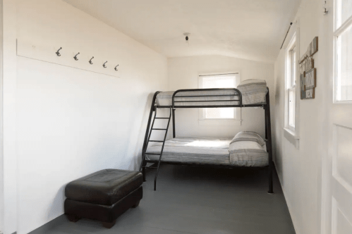 A simple bedroom featuring a bunk bed, a brown ottoman, and hooks on the wall, with natural light from windows.