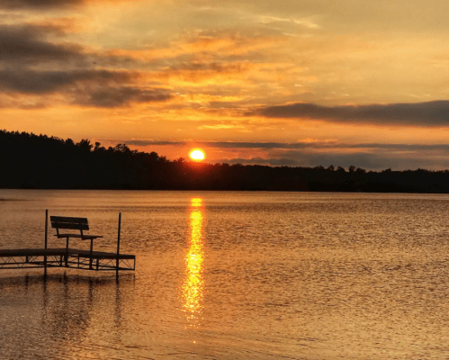 A serene lake at sunset, with a bench on a dock reflecting the orange and gold hues in the water.