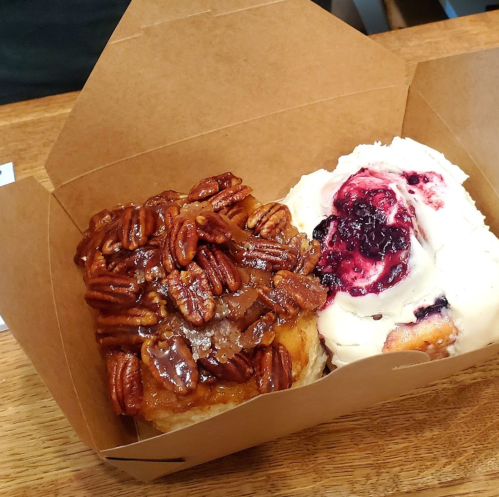 A takeout box containing a pecan-topped pastry and a scoop of ice cream with berry sauce.