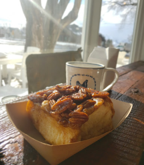 A warm cinnamon roll topped with pecans, served on a wooden table next to a coffee cup, with a snowy view outside.
