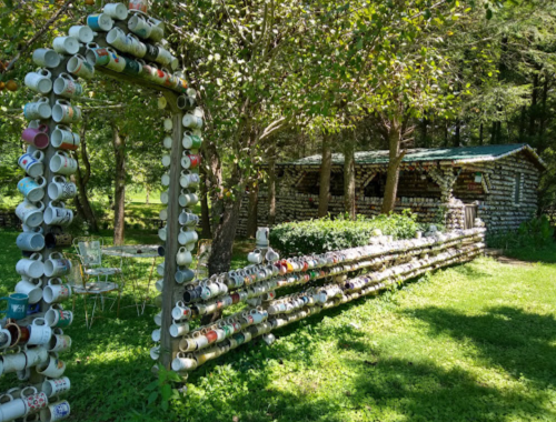 A whimsical outdoor scene featuring a fence and archway made of colorful mugs, with a rustic cabin in the background.