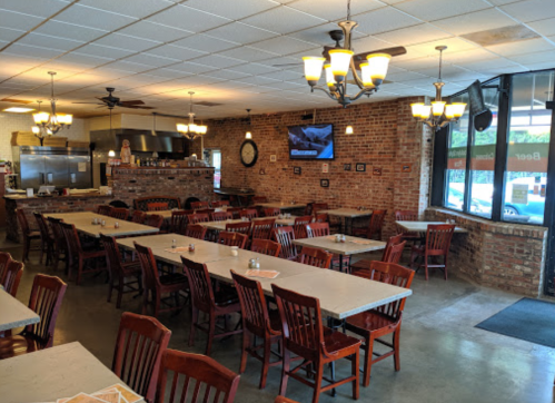 Interior of a restaurant featuring wooden tables and chairs, brick walls, and large windows with natural light.
