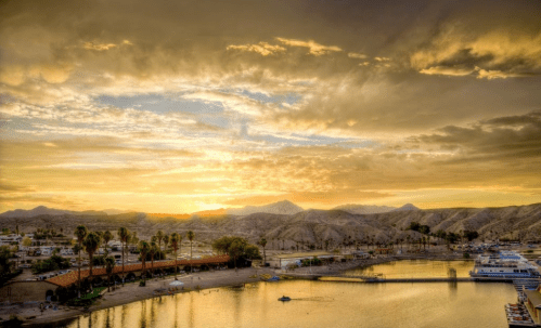 Sunset over a serene lake surrounded by mountains, with palm trees and a sandy beach in the foreground.