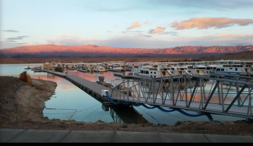 A serene marina at sunset, with boats docked and mountains in the background reflecting warm colors on the water.
