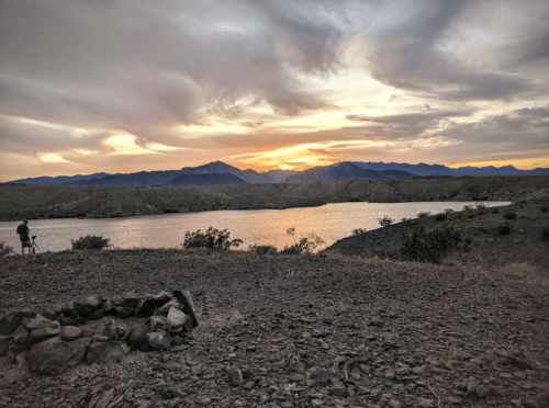 A serene sunset over a lake, framed by mountains and rocky terrain, with a person capturing the view in the foreground.