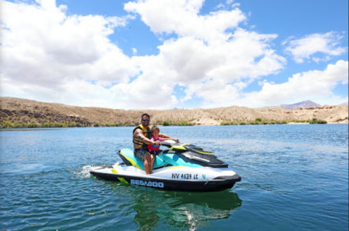 A person and a child ride a jet ski on a lake, surrounded by mountains and a blue sky with fluffy clouds.