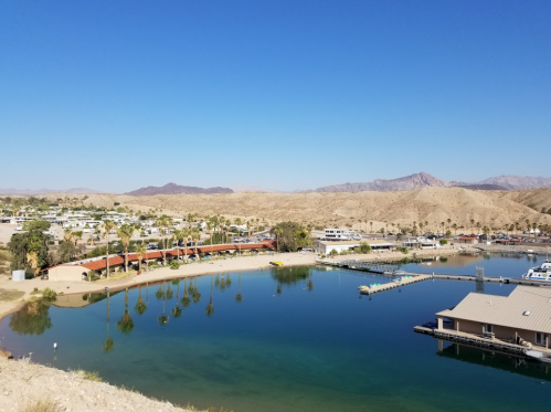 A serene marina surrounded by desert hills under a clear blue sky, with boats docked and palm trees lining the shore.
