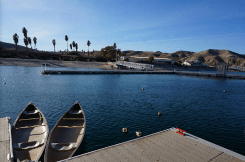 Two canoes on a dock by a calm lake, with palm trees and hills in the background under a clear blue sky.