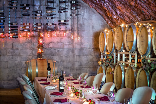 A beautifully set dining table in a wine cellar, surrounded by wine barrels and soft lighting.