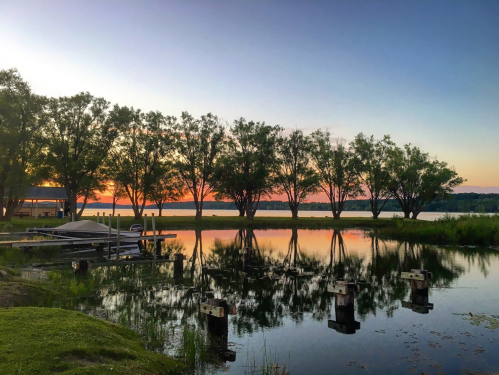 A serene lake at sunset, with trees reflecting on the water and a dock in the foreground.