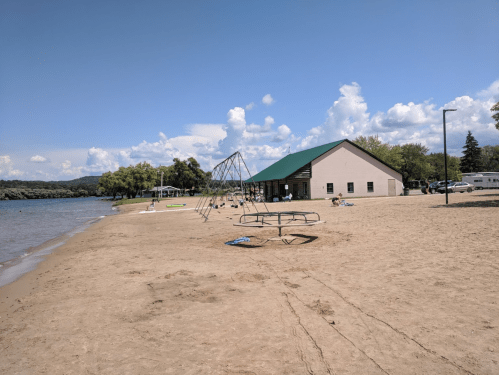 A sandy beach with a playground, a building, and people enjoying the sunny day by the water.