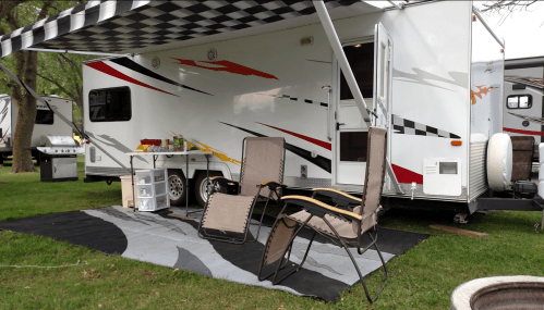 A camper with a checkered awning, two chairs, and a small table set up on a grassy area.