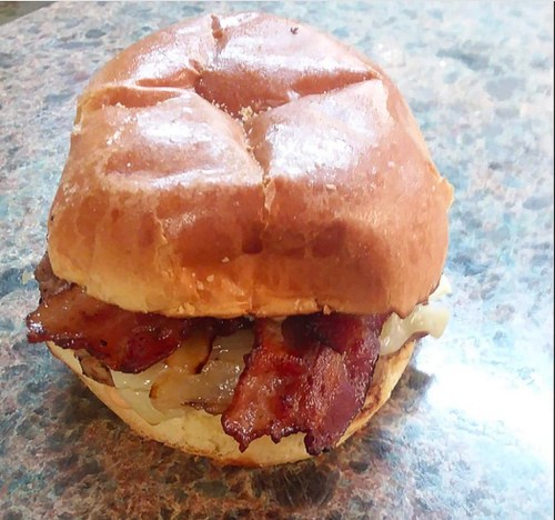 A close-up of a bacon sandwich on a soft bun, with crispy bacon and sautéed onions, resting on a textured surface.