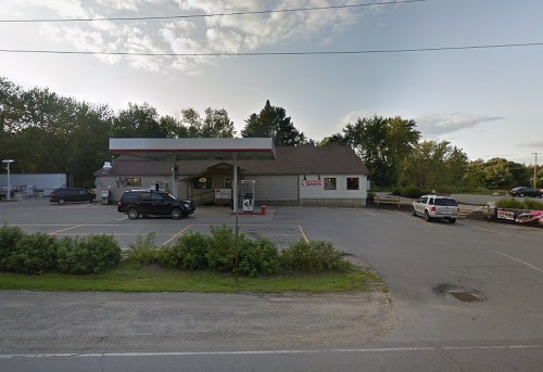 A small gas station with a convenience store, surrounded by trees and parked cars, under a clear sky.
