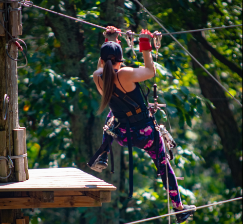 A person in athletic gear zip-lining through a forest, gripping the harness with one hand and legs extended.