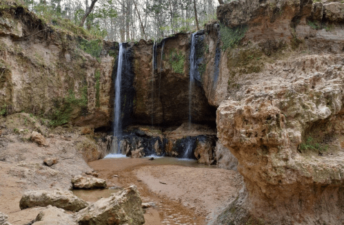 A serene landscape featuring two waterfalls cascading over rocky cliffs into a sandy riverbed surrounded by greenery.