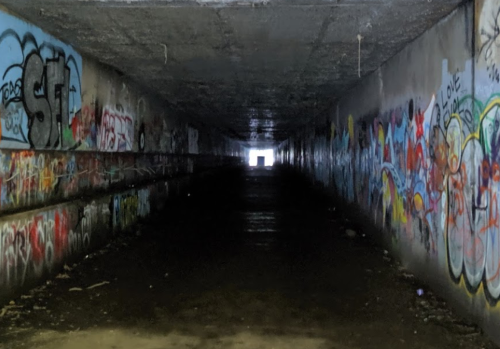 A dark tunnel with colorful graffiti on the walls, leading to a bright opening at the far end.