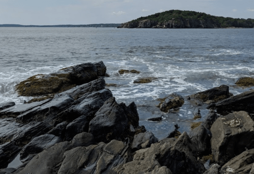 Rocky shoreline with waves crashing, leading to a distant green island under a clear sky.