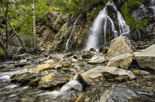 A serene waterfall cascades over rocks, surrounded by lush greenery and flowing water in a tranquil natural setting.