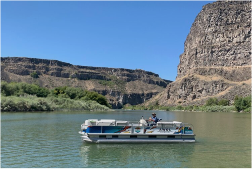A person on a pontoon boat named "AWOL" floats on a calm river surrounded by rocky cliffs and greenery.