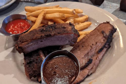 A plate with two slices of smoked brisket, fries, and small bowls of barbecue sauce and ketchup.