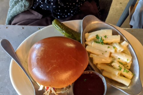 A plate with a burger, a pickle, and creamy pasta on the side, served on a metal table.