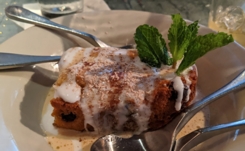 A slice of dessert topped with cream, mint leaves, and a sprinkle of cinnamon, served on a white plate with spoons.